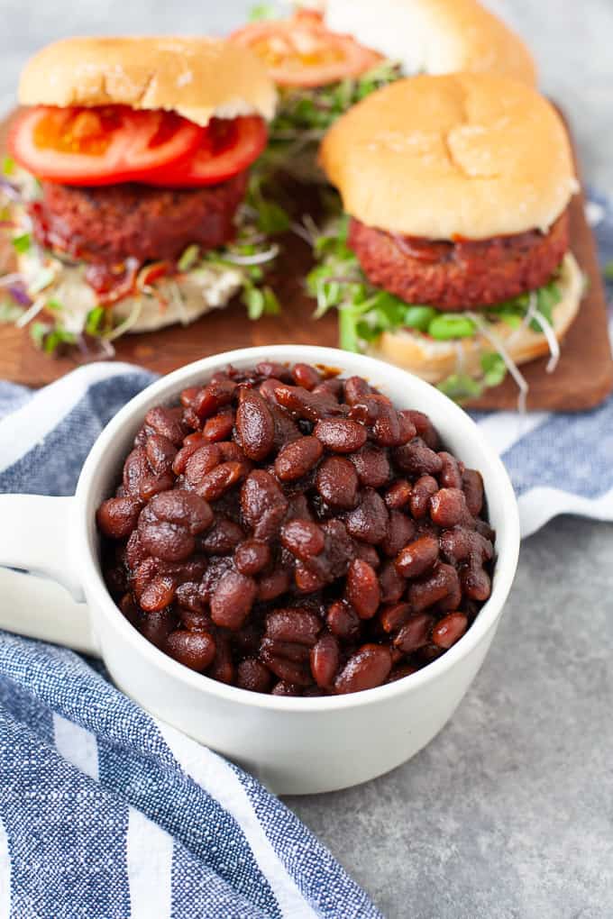 baked beans. in white mug. striped napkin. two loaded burgers. on cutting board.